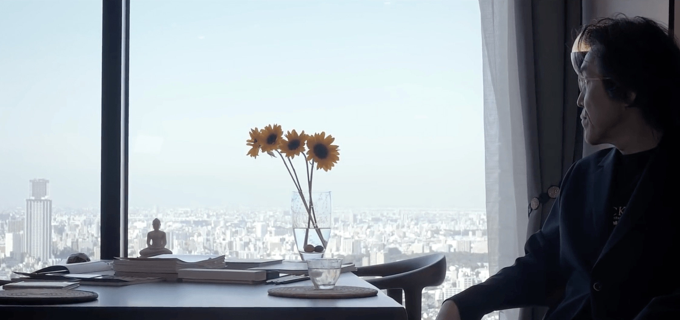 Miyamoto sitting by a window above the city, notebooks and a vase of sunflowers on the table