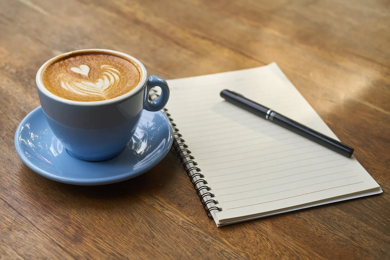 A cup of coffee beside an open blank notebook and a pen on a wooden table
