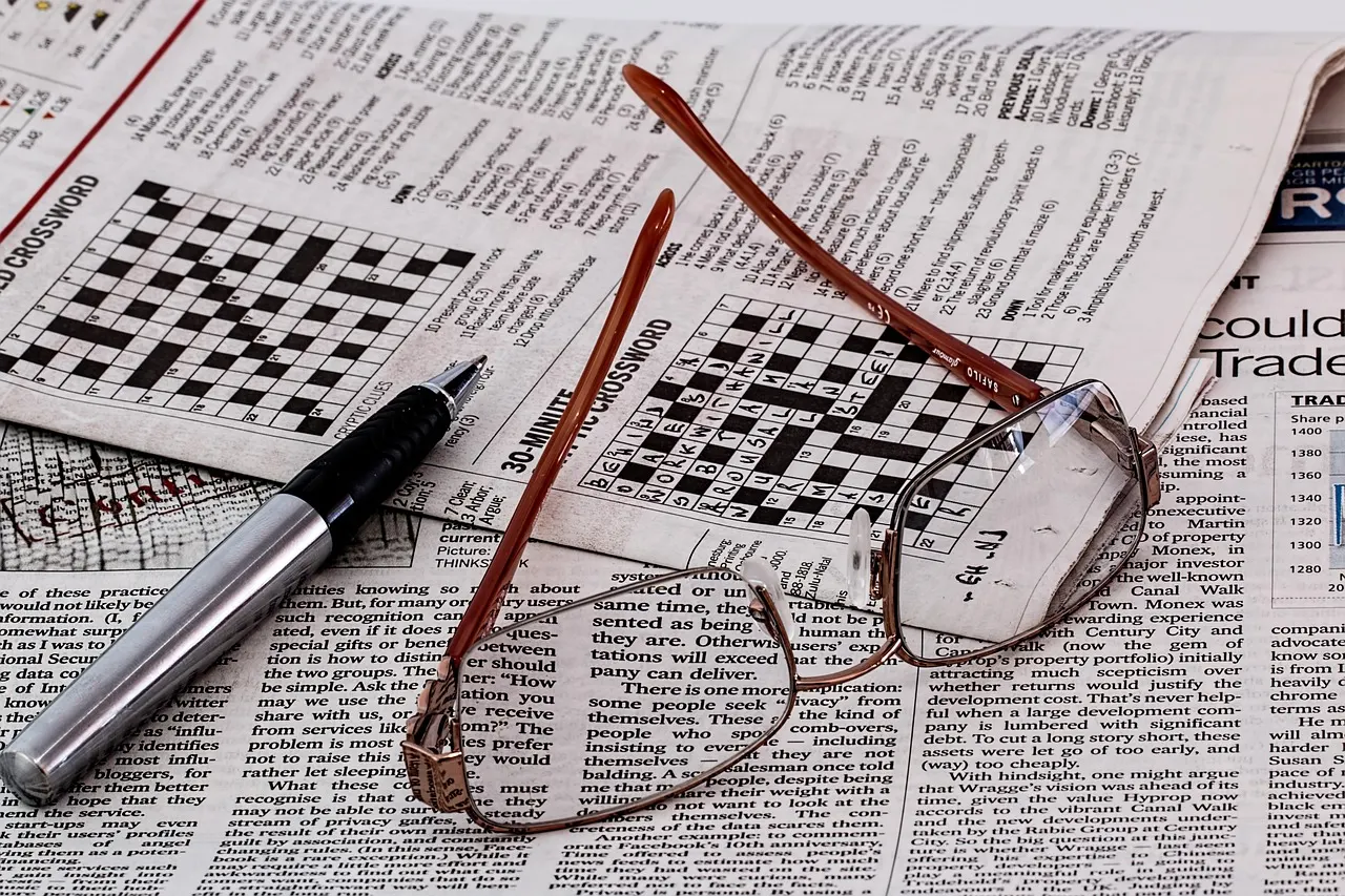 A newspaper folded open at a crossword, with a pen and reading glasses resting on it
