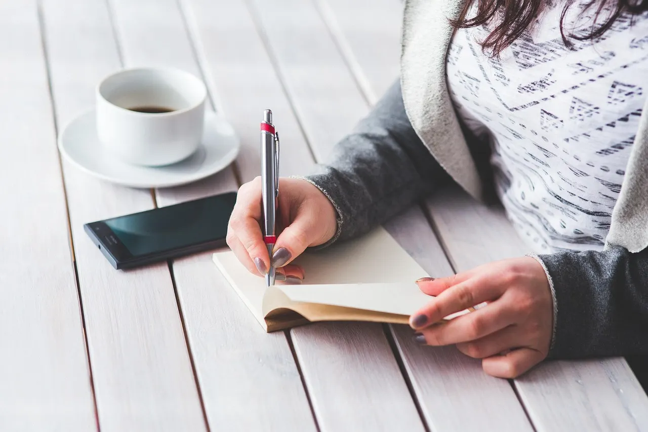 A woman writing in a notepad beside a cup of coffee at a table in daylight