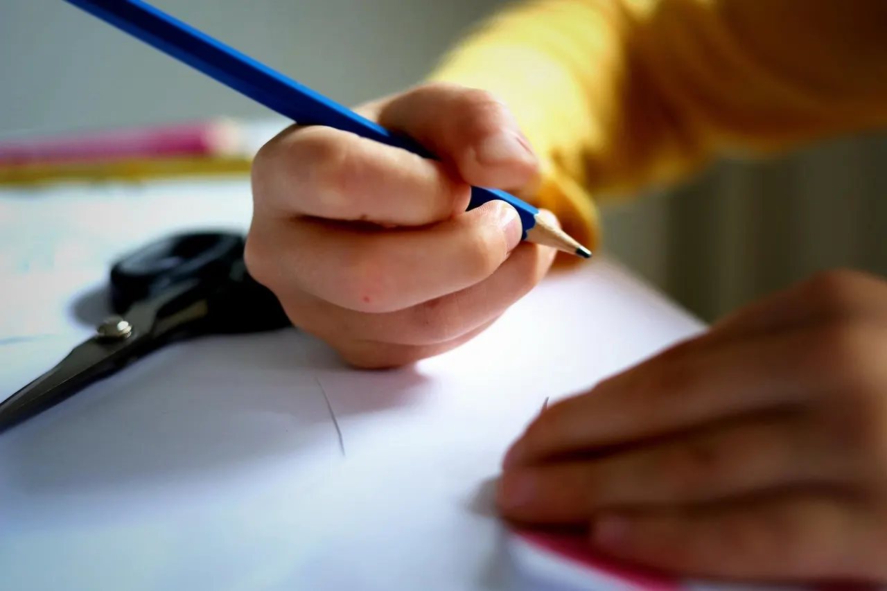 A child's hand holding a pencil, writing on a sheet of paper at a desk